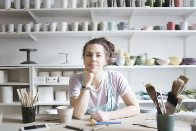 Portrait of confident young female potter sitting at workbench against shelves at store
