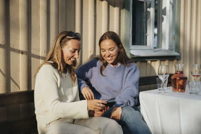 Happy young woman sharing smart phone with female friend during dinner party at cafe