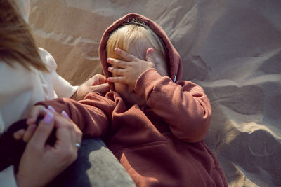 Boy in a hoodie covers his face with his hand as he lies on his back on the sand next to his mother