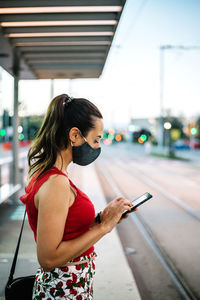 Side view of young female in black protective mask browsing smartphone while standing on railway platform and waiting for train