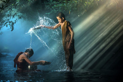 Boy wearing traditional clothing while pouring water on teenager in lake at forest