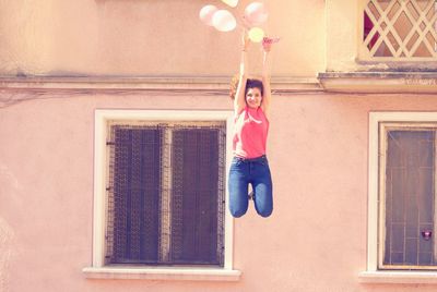 Smiling young woman holding balloons while jumping against building