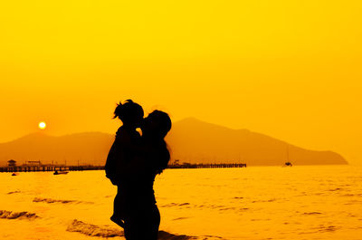 Silhouette woman standing with daughter at beach against orange sky