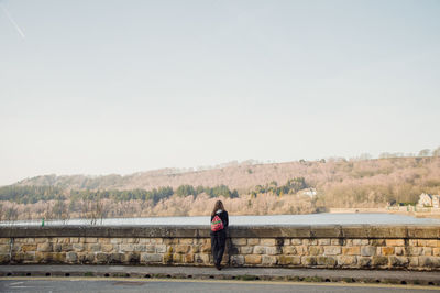 Rear view of woman standing on retaining wall by river against clear sky