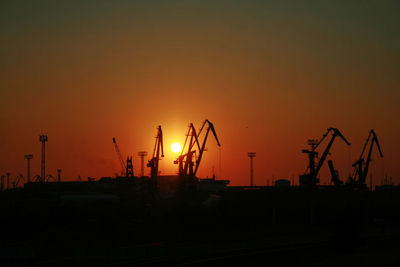 Silhouette cranes at construction site against sky during sunset