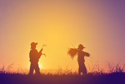 Teenage boy holding sickles while girl carrying crop on field against sky during sunset