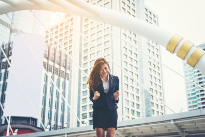 Happy young businesswoman clenching fists against buildings in city