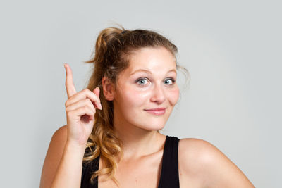 Close-up portrait of smiling young woman pointing upwards against gray background