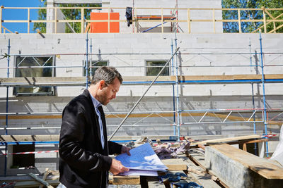 Side view of architect reading blueprint by incomplete building at construction site