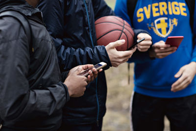 Midsection of phone addicted friends wearing warm clothing while standing on basketball court