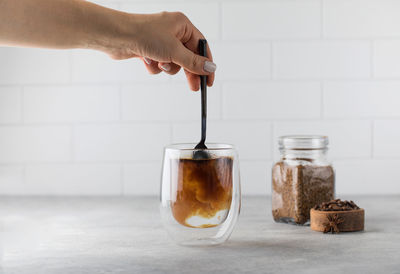 Woman stirs instant coffee in glass mug with boiled water on grey stone table