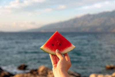 Personal perspective of hand holing piece of watermelon on beach.