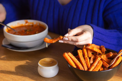 Woman hand dips a slice of sweet potato fries in sweet chili sauce, eating them in cafe, restaurant.