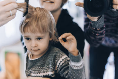 Midsection of man photographing barber cutting girl's hair sitting with mother in salon