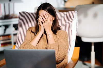 Unrecognizable female sitting in comfortable armchair and hiding face behind hands in embarrassment while having fun at home