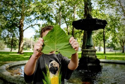 Boy hiding face with leaf against fountain at park