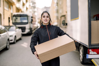 Portrait of young delivery woman carrying box while standing on street in city