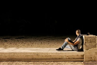 Side view of mid adult man sitting on retaining wall against sky at night
