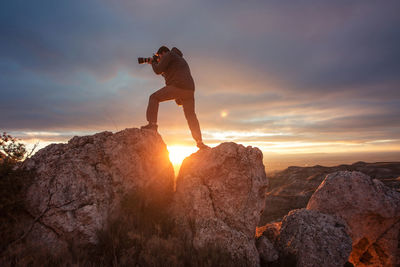 Man photographing while standing on rock against sky during sunset