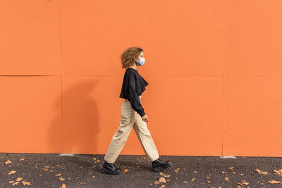 Young woman wearing face mask walking against orange wall