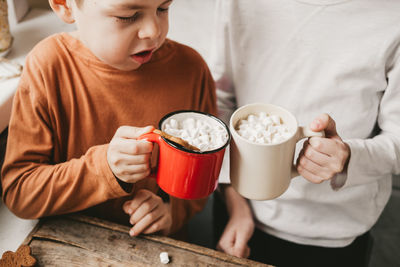 Two boy in christmas clothes holding cocoa with marshmallows in the kitchen.