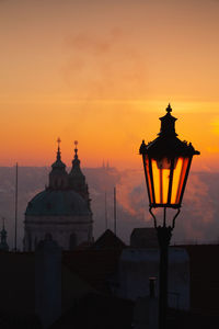 Illuminated street light by building against sky during sunset