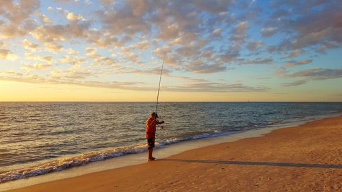 Man holding fishing rod while standing at beach against sky during sunset