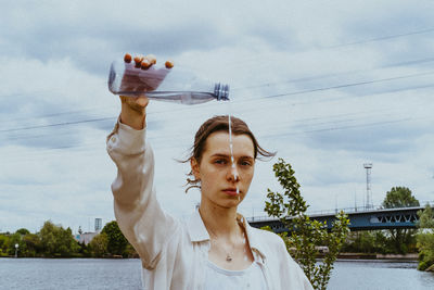 Portrait of confident young non-binary person pouring water from bottle against sky