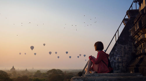 Side view of woman looking at hot air balloons flying against clear sky during sunset