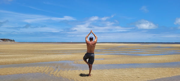 Man practices yoga position on the sand in front of the sea with blue sky
