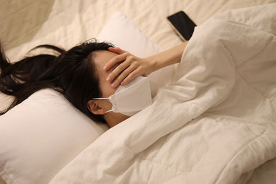 Sick woman who wearing a mask lying in bed, her hands over her eyes, girl sleeping in bed