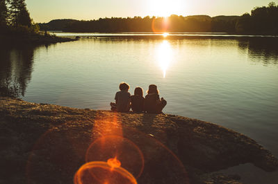 Children sitting by lake against sky during sunset