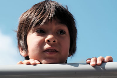 Close-up of boy looking away while leaning on railing against blue sky