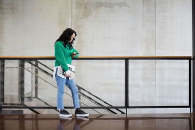 Young female student using mobile phone while standing at railing against wall in university