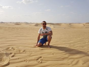 Portrait of man kneeling on sand in desert against sky