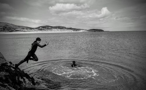 Side view of boy jumping in sea against cloudy sky