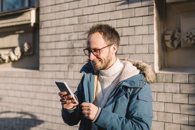 Handsome male with beard, glasses and in blue jacket looks the way to his hotel on smartphone. 