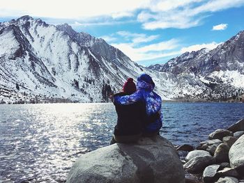 Friends with arm around sitting at lakeshore against snowy mountains