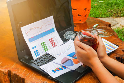 Cropped hand of woman holding tea at table with laptop and documents