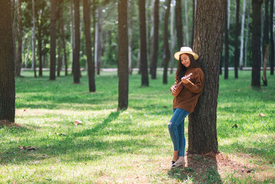 Woman playing ukulele while standing against tree trunk