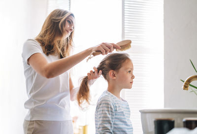 Young woman with little tween girl daughter in pajamas combs her hair  in bathroom at home