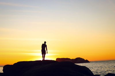 Rear view of silhouette man standing at beach against sky during sunset