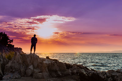 Man standing on rock by sea against sky during sunset