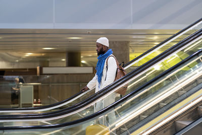 African american passenger man with suitcase stands on escalator, holds handrail in airport terminal