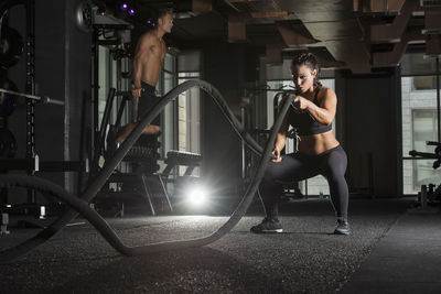 Young woman exercising with battle ropes while man in background at gym