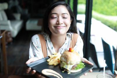 Close-up of smiling young woman with eyes closed holding fast food in restaurant