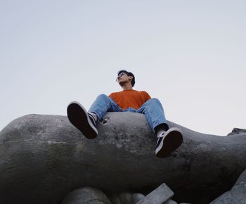 Low angle view of man sitting on rock against clear sky