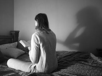 Rear view of woman reading book on bed at home