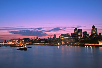 Illuminated buildings by river against sky at sunset