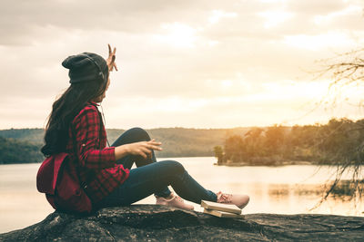 Side view of young woman sitting by lake against sky